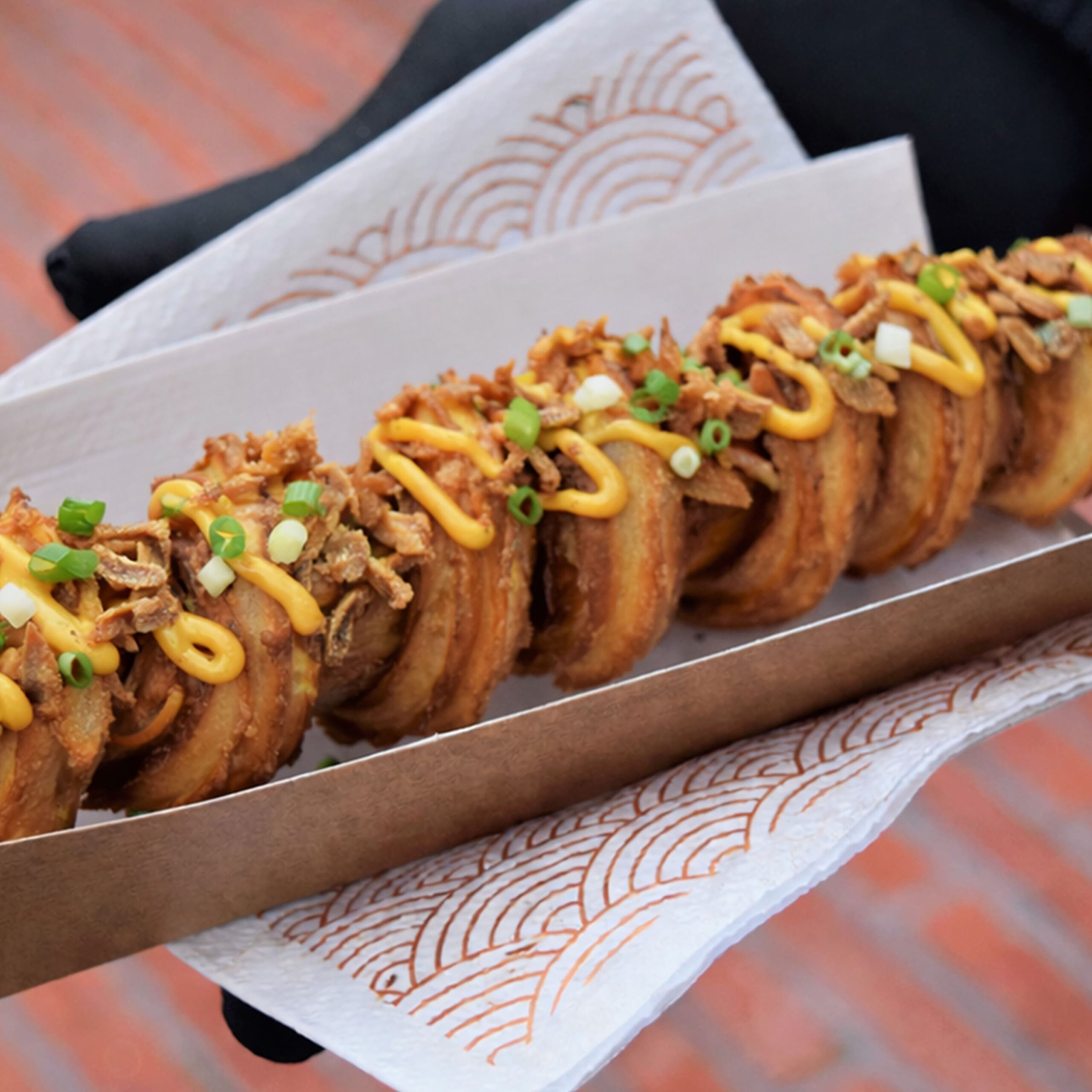 photo de notre Churrosdog artisanal croustillant nappé de sauce onctueuse et parsemé d'oignons frits et cébettes présenté dans un emballage kraft dans une ambiance street food