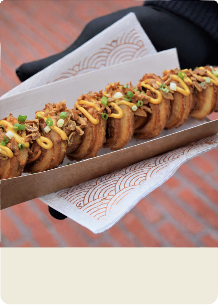 photo de notre Churrosdog artisanal croustillant nappé de sauce onctueuse et parsemé d'oignons frits et cébettes présenté dans un emballage kraft dans une ambiance street food