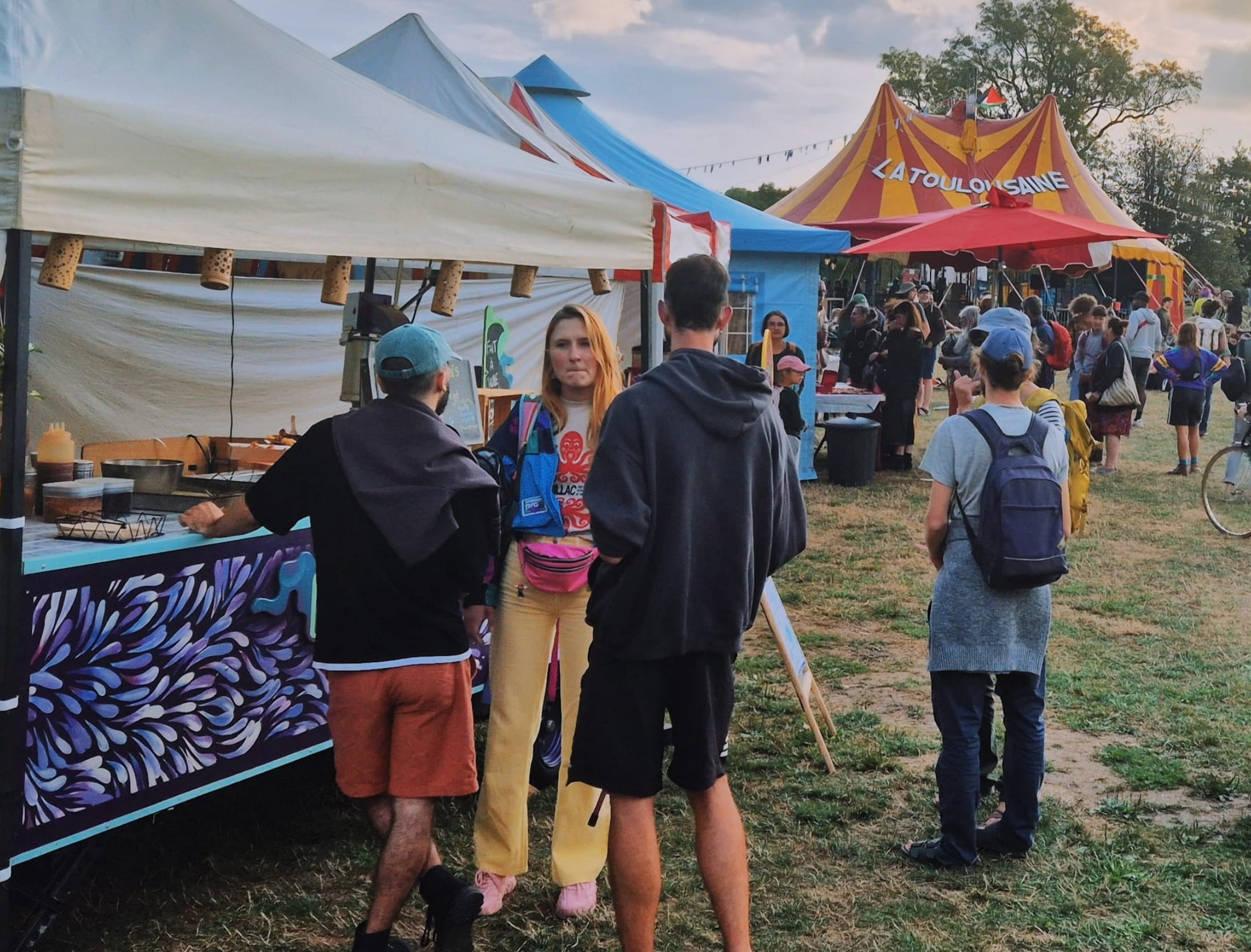 Ambiance de festival rétro avec le stand Churringa et ses motifs colorés, situé à côté d'un chapiteau 'La Toulousaine' dans un champ verdoyant.